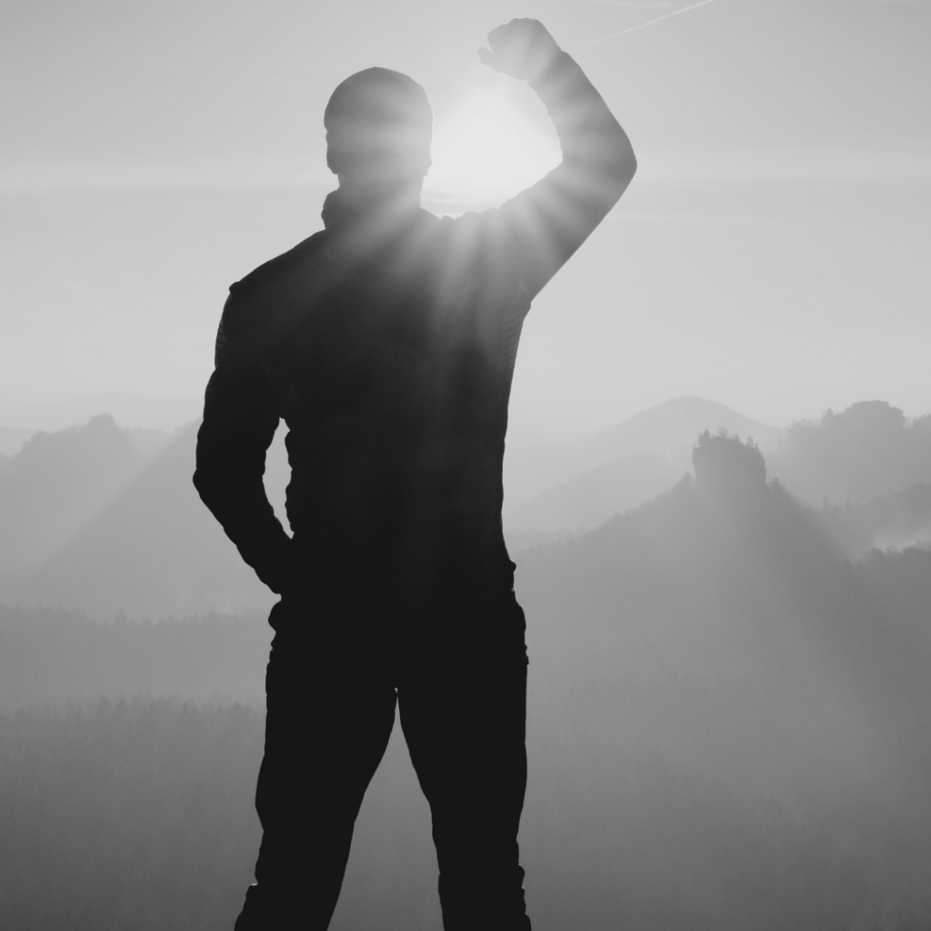 Silhouette eines Mannes mit erhobenem Arm gegen die aufgehende Sonne in einer Berglandschaft.